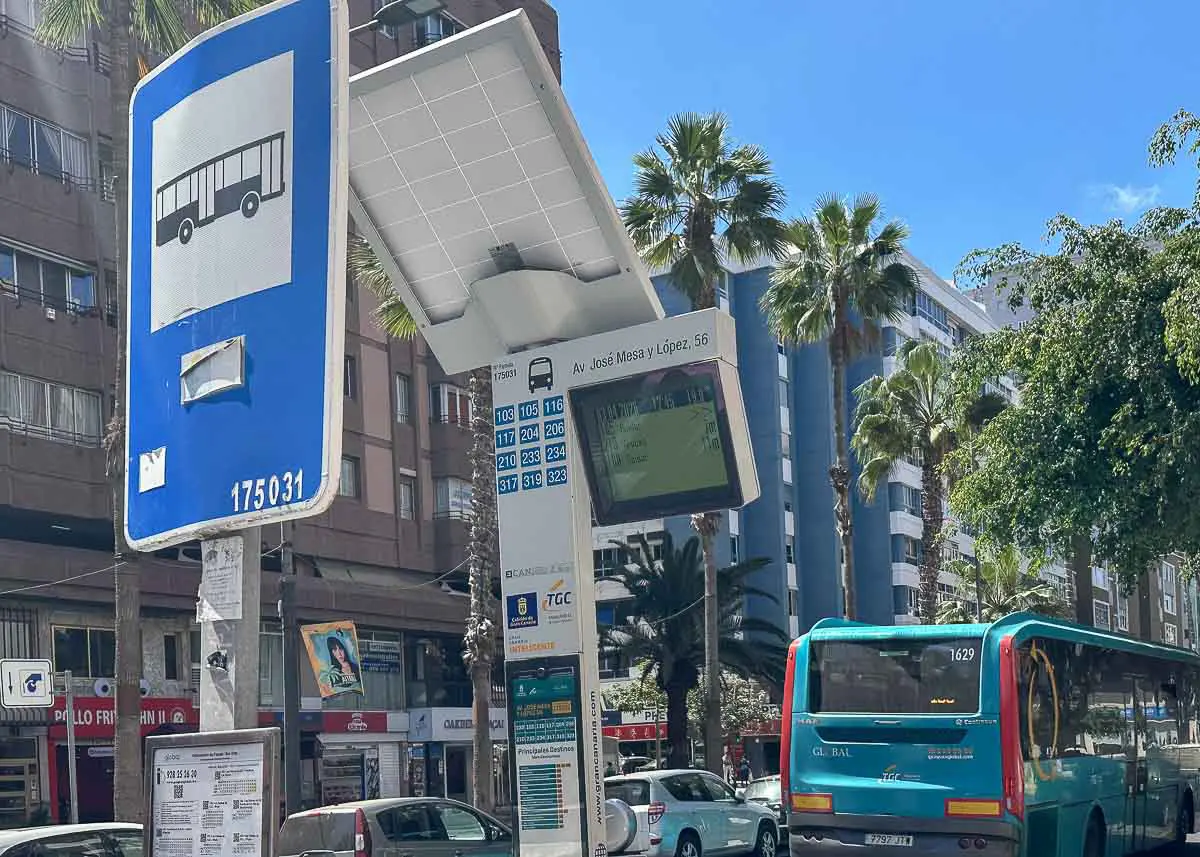 Eine Bushaltestelle in Las Palmas de Gran Canaria mit einer digitalen Anzeigetafel, einem großen Bussymbol, einem geparkten blauen Bus, Palmen und hohen Gebäuden im Hintergrund an einem sonnigen Tag.