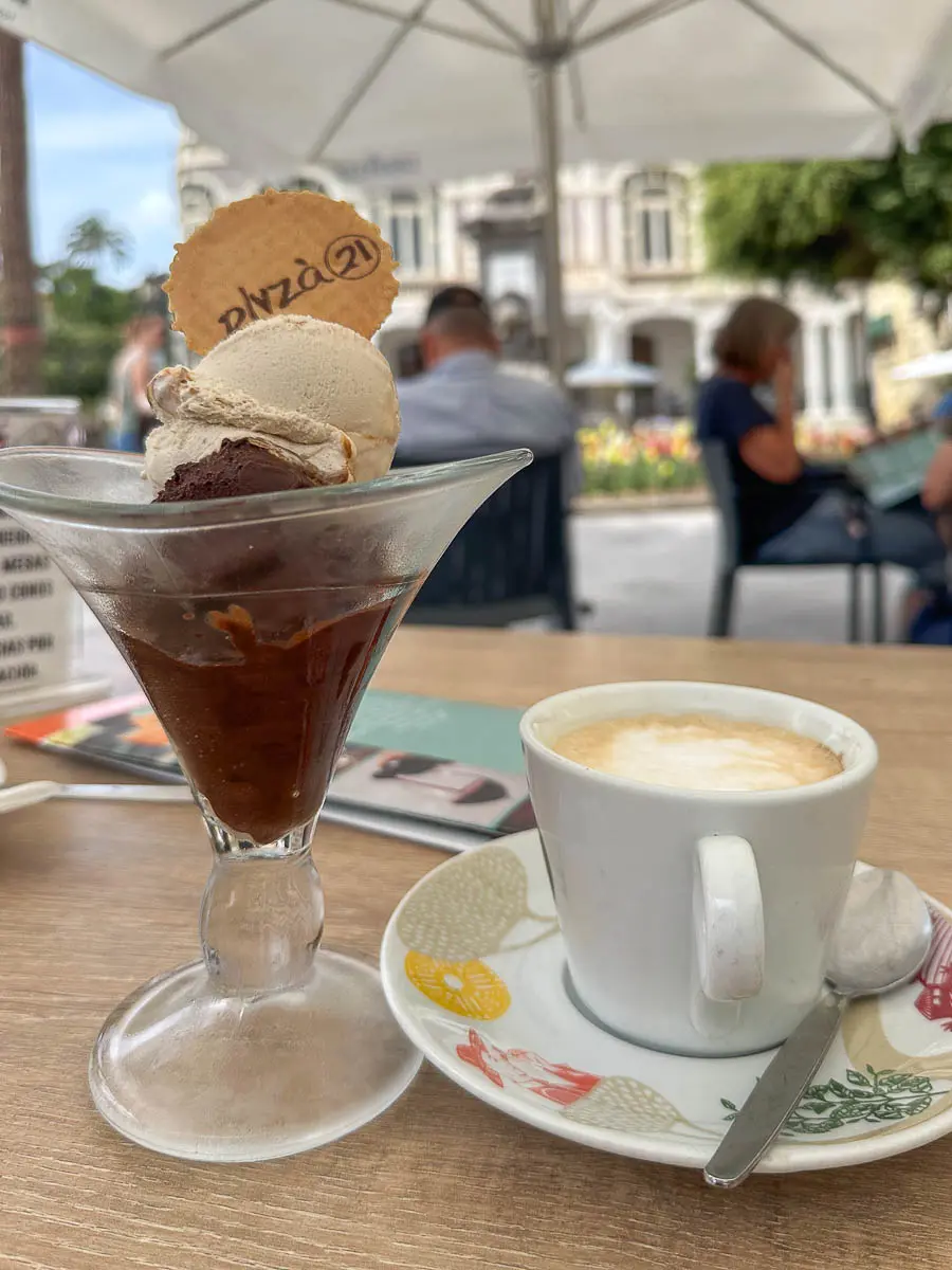 Auf einem Tisch im Freien eines Cafés in Las Palmas de Gran Canaria steht ein Glas mit Eis und einer Waffel neben einer Tasse Cappuccino auf einer Untertasse, im Hintergrund sind Menschen und Sonnenschirme zu sehen – ideal, um sich nach dem Erkunden der örtlichen Sehenswürdigkeiten zu entspannen oder Tipps von anderen Reisenden zu erhalten.