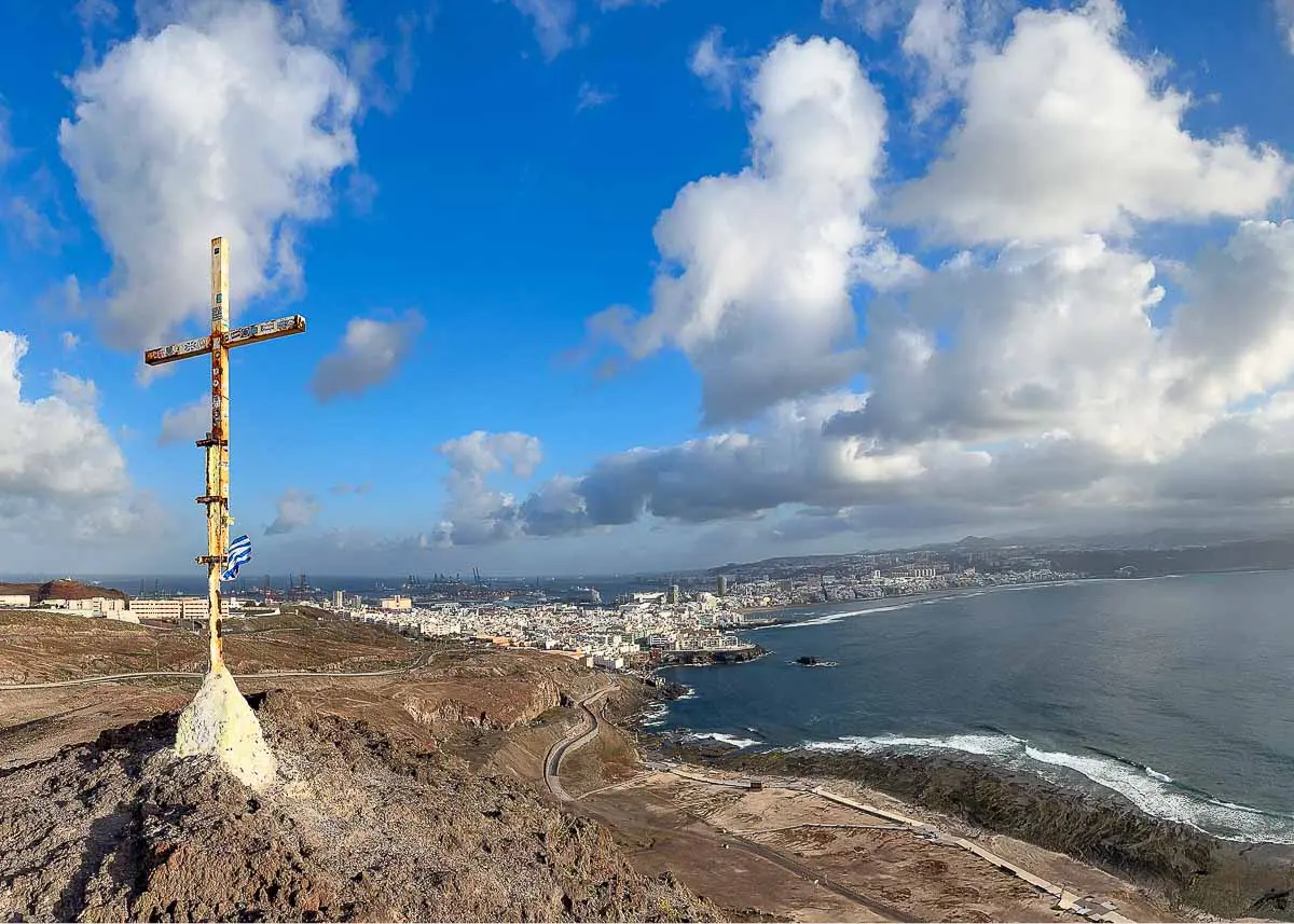 Auf einem Hügel mit Blick auf Las Palmas de Gran Canaria steht ein verwittertes Kreuz. Die Küstenstadt liegt unter strahlend blauem Himmel mit vereinzelten Wolken, und in der Ferne sind das Meer und die Küste zu sehen – einer der besonderen Tipps für Ihren Besuch.