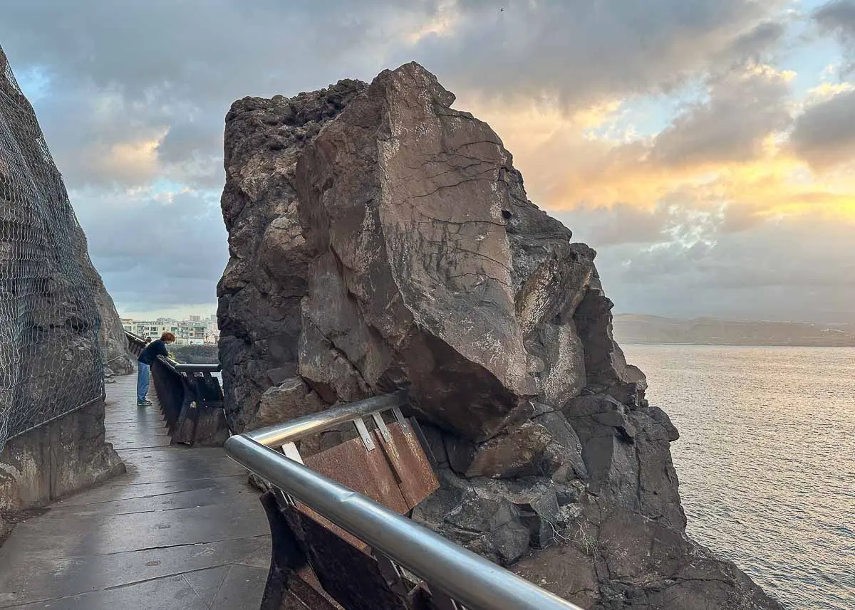 Eine Person steht bei Sonnenuntergang auf einem Küstenweg in der Nähe einer Felsformation an der Playa Confital, im Hintergrund sind das Meer und der bewölkte Himmel zu sehen, und entlang des Weges verlaufen Metallgeländer – einer der Lieblingsorte, die Las Palmas de Gran Canaria so charmant machen.
