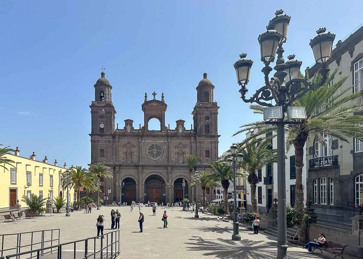Am Ende eines sonnigen, von Palmen gesäumten Platzes erhebt sich eine historische Kathedrale mit zwei Türmen, die zu den wichtigsten Sehenswürdigkeiten in Las Palmas de Gran Canaria zählt. Auf dem weitläufigen Platz, der von klassischen Straßenlaternen und farbenfrohen Gebäuden umgeben ist, flanieren Menschen und treffen sich – ein idealer Ort für Sightseeing-Tipps.