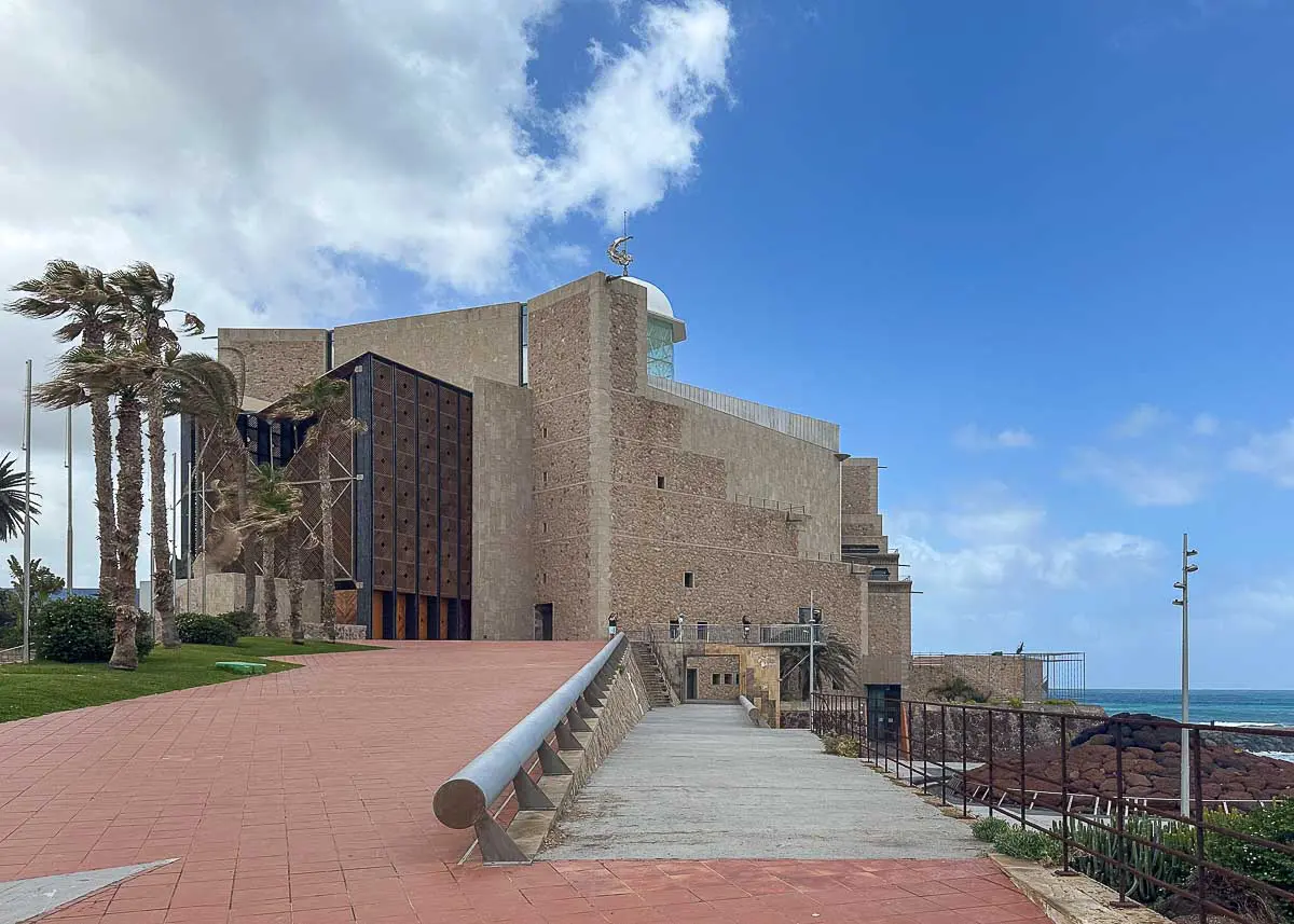 Ein modernes Steingebäude mit geometrischen Formen in der Nähe des Meeres in Las Palmas de Gran Canaria, Palmen, die sich im Wind wiegen, und ein teilweise bewölkter Himmel darüber. Ein mit roten Fliesen gepflasterter und ein betonierter Weg führen zum Eingang und machen das Auditorio Alfredo Kraus zu einer der Top-Sehenswürdigkeiten.