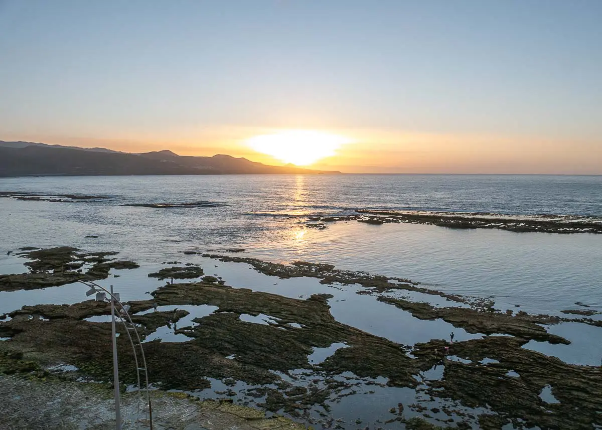 Sonnenuntergang über einem ruhigen Meer mit felsigen Gezeitentümpeln an der Playa de las Canteras im Vordergrund und fernen Bergen am Horizont unter einem klaren Himmel