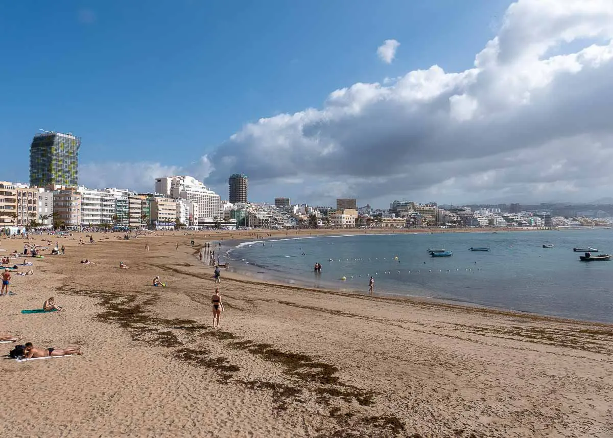 Am breiten Sandstrand Playa Las Canteras in Las Palmas de Gran Canaria entspannen sich die Menschen, im Hintergrund sind Gebäude und Hotels zu sehen. In der Bucht herrscht ruhiges Wasser, und kleine Boote sind zu sehen. Der Himmel ist teilweise bewölkt und wirft sanfte Schatten auf den Strand und die Stadt.