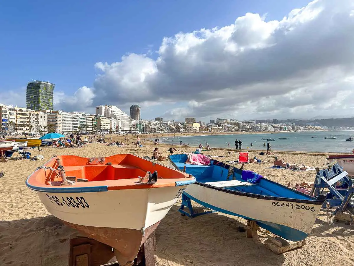 Am Sandstrand Playa de las Canteras in Las Palmas de Gran Canaria liegen bunte Boote, während sich Menschen sonnen und spazieren gehen. Im Hintergrund sind das Meer, hohe Gebäude und ein bewölkter blauer Himmel zu sehen