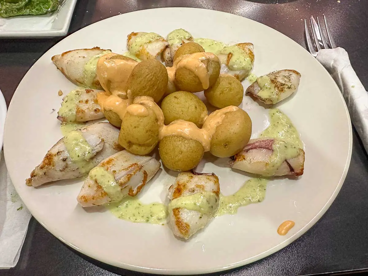 Ein Teller mit gegrilltem Tintenfisch und kleinen runden Kartoffeln, übergossen mit grüner und orangefarbener Soße, serviert auf einem weißen Teller mit einer Gabel und einer Serviette daneben – eine köstliche Art, die lokalen Aromen zu genießen, während man die Sehenswürdigkeiten von Las Palmas de Gran Canaria erkundet.