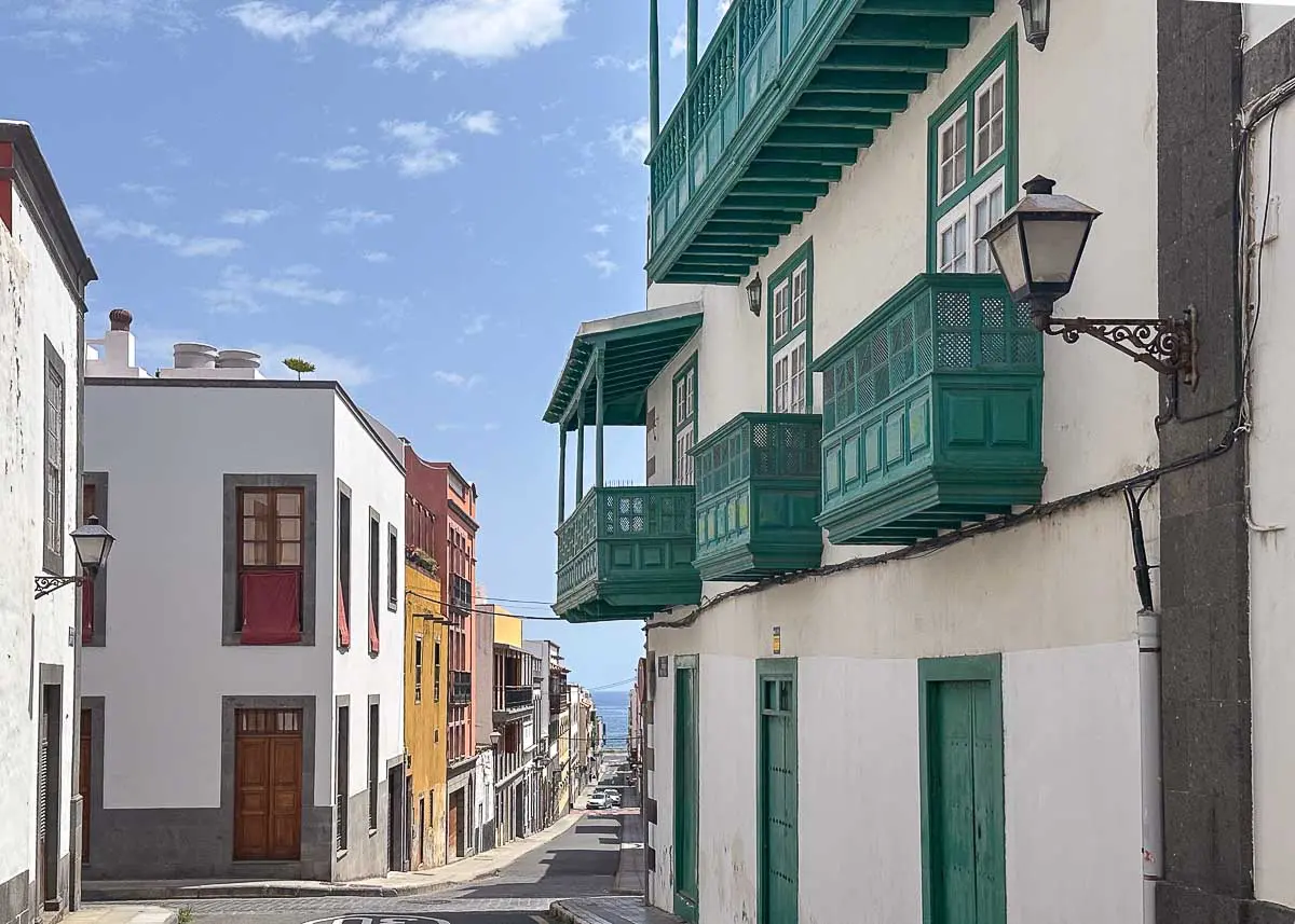 Eine schmale Straße in einer mediterranen Stadt mit weißen Gebäuden und grünen Holzbalkonen unter einem blauen Himmel mit vereinzelten Wolken. Die Straße führt bergab in Richtung des in der Ferne liegenden Meeres – eine Szene, die an die charmanten Viertel und versteckten Sehenswürdigkeiten in Las Palmas de Gran Canaria erinnert.