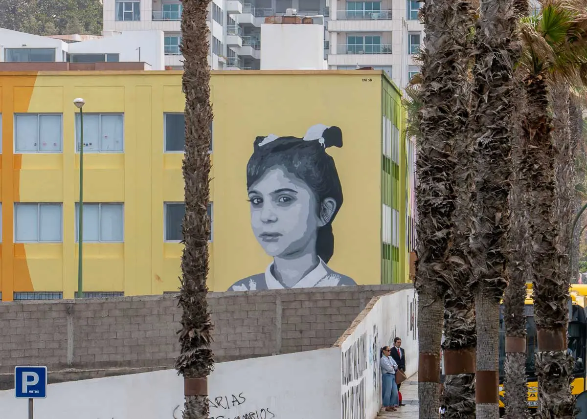 An einem gelben Gebäude hinter Palmen in Las Palmas de Gran Canaria ist ein großes Schwarz-Weiß-Wandbild eines Mädchens mit Zöpfen zu sehen. In der Nähe steht eine Gruppe von drei Personen an einer Mauer, und im Vordergrund ist ein blaues Parkschild zu sehen – eine der auffälligen Sehenswürdigkeiten, die sich hervorragend als lokale Tipps für die Erkundung der Stadt eignen.
