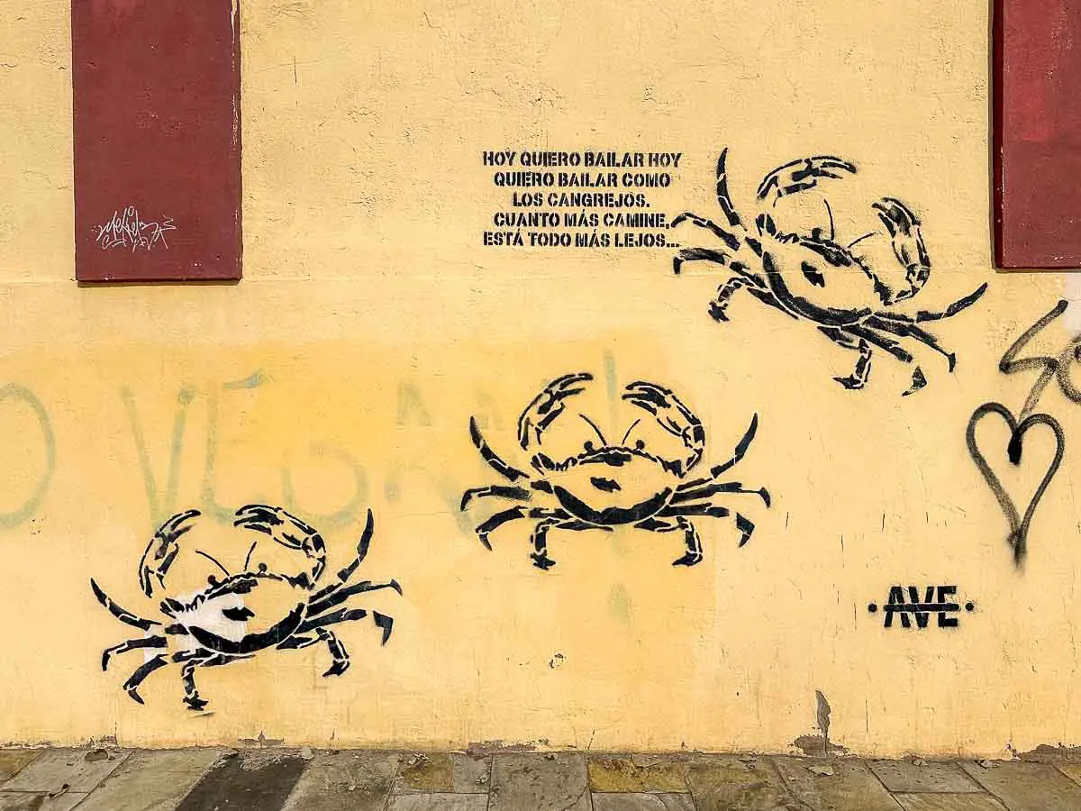Auf einer gelben, verwitterten Wand mit roten Akzenten, verblassten Graffiti-Spuren und einem Herzsymbol auf der rechten Seite sind drei schwarze Krabben im Schablonentechnik sowie ein Graffiti-Schriftzug auf Spanisch zu sehen. Dieser auffällige Ort in Las Palmas de Gran Canaria gehört zu den versteckten Sehenswürdigkeiten, die neugierige Besucher anziehen. Der Bürgersteig besteht aus quadratischen Steinen – ein großartiger Ort für einzigartige Reisetipps oder unvergessliche Fotos.