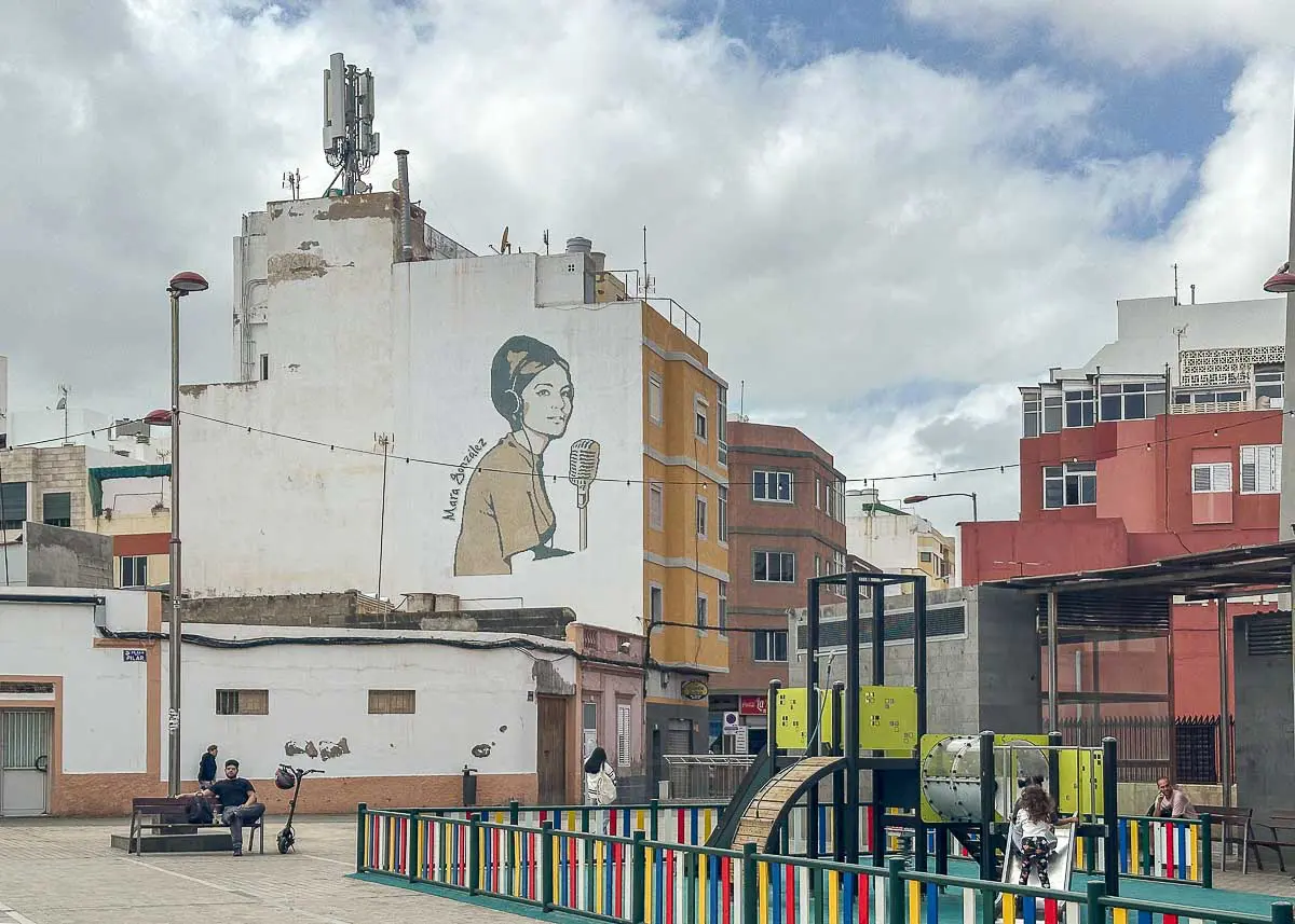 Vor einigen Gebäuden in Las Palmas de Gran Canaria befindet sich ein Spielplatz mit bunten Zäunen. An einem der Gebäude ist ein großes Wandgemälde zu sehen, das eine Frau zeigt, die in ein Mikrofon singt – eine der einzigartigen Sehenswürdigkeiten der Gegend. Überall in der Gegend sind Menschen zu sehen, die trotz des bewölkten Himmels die lebhafte Atmosphäre genießen – ideal für alle, die auf der Suche nach Insidertipps zur Erkundung der lokalen Kunst und Kultur sind.
