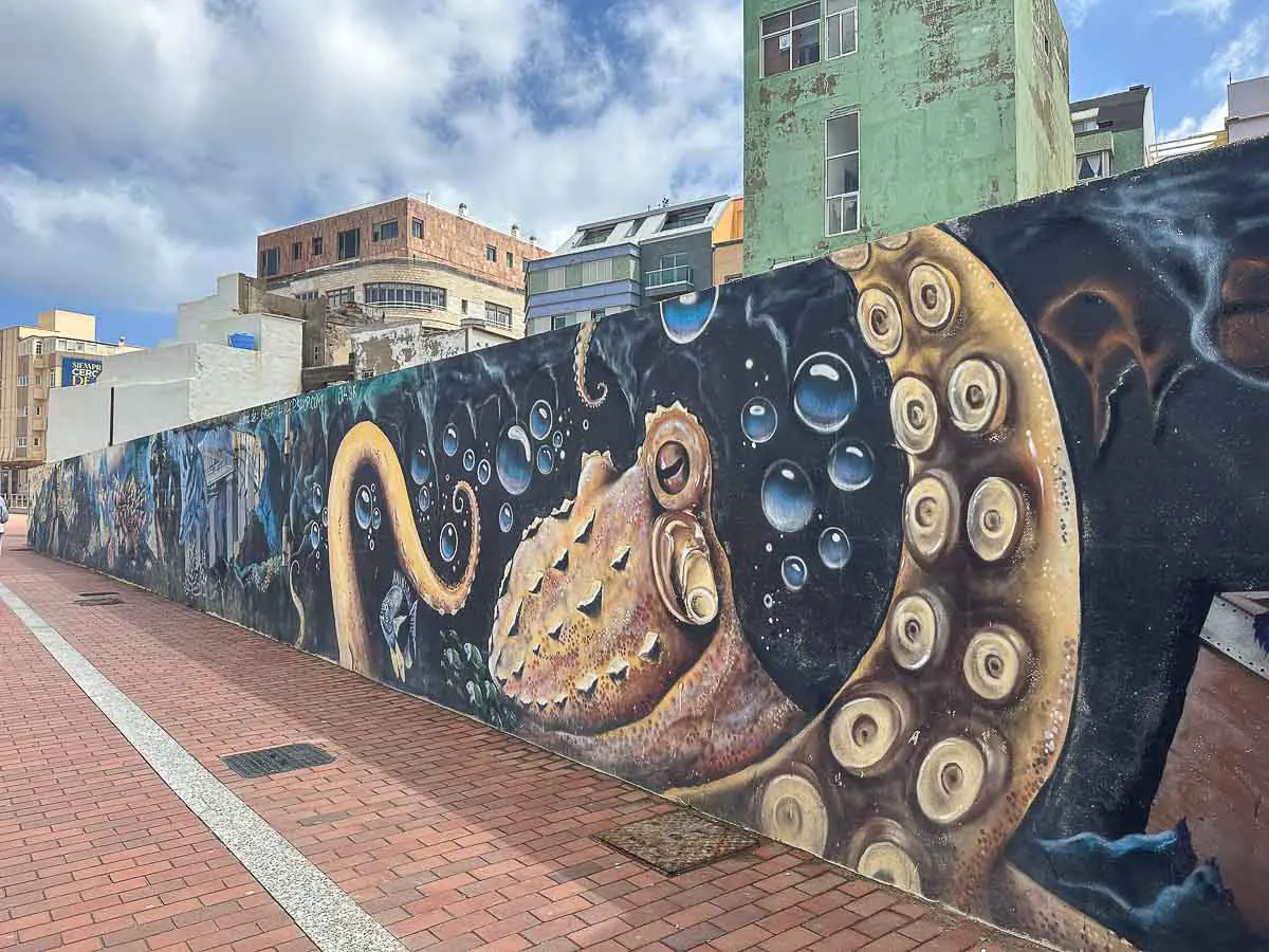 Ein farbenfrohes Wandgemälde an einer Stadtmauer in Las Palmas de Gran Canaria zeigt einen großen Tintenfischarm, Blasen und Unterwasserlebewesen vor dem Hintergrund städtischer Gebäude unter einem teilweise bewölkten Himmel – eine der einzigartigen Sehenswürdigkeiten der Stadt und ideal für Tipps zur Entdeckung ihrer lebendigen Straßenkunst.