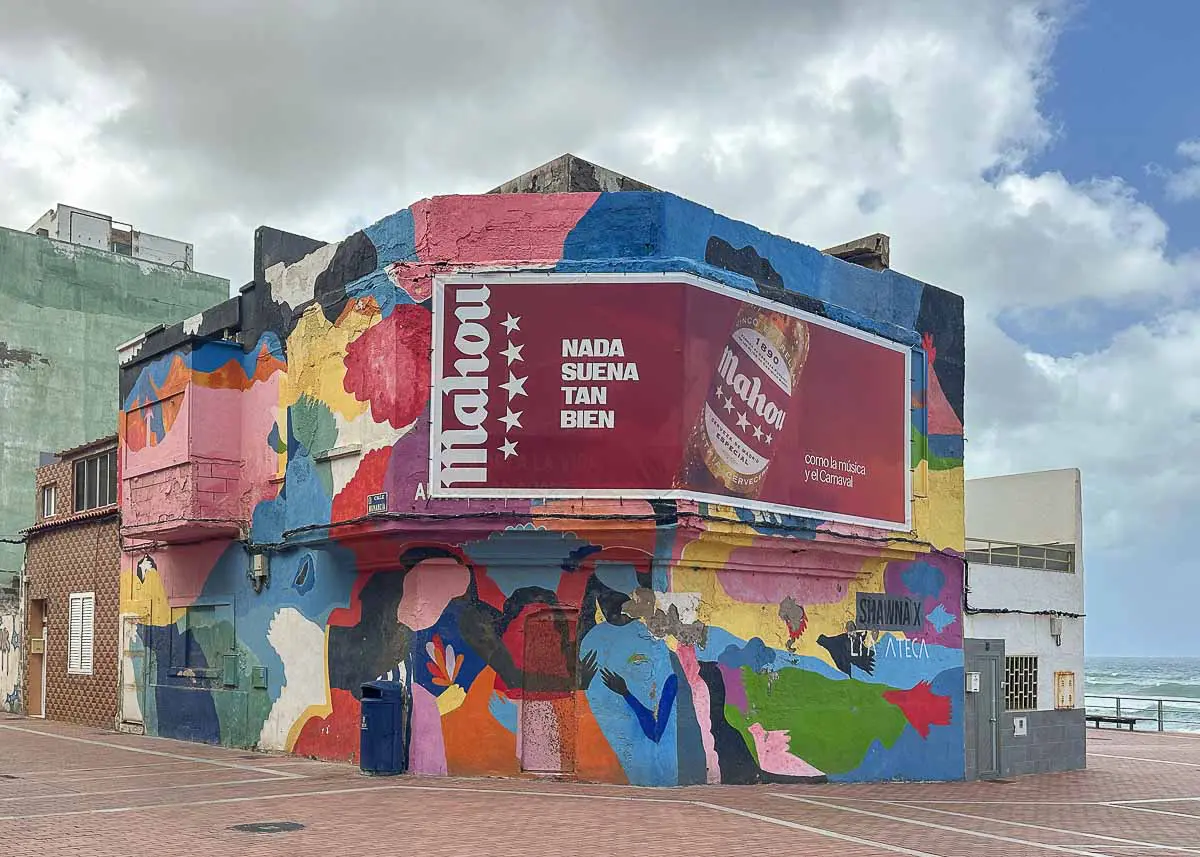 Die Casa de Color ist ein farbenfrohes, abstrakt mit Streetart bemaltes Gebäude am Meer in Las Palmas de Gran Canaria. Es trägt eine große rote Werbetafel für Mahou-Bier mit dem Slogan „Nada suena tan bien“. Der Himmel ist bewölkt und die Straße ist leer.