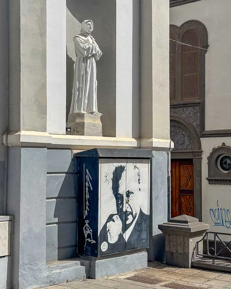Über einem bemalten Versorgungskasten mit einem Schwarz-Weiß-Porträt einer Person, die ein Kind im Arm hält, thront eine weiße Statue einer in eine Robe gekleideten Figur vor dem Hintergrund einer grauen Gebäudefassade mit Holztüren und verzierten Fenstern – eine der weniger bekannten Sehenswürdigkeiten in Las Palmas de Gran Canaria und ein idealer Ort für einzigartige Tipps bei der Erkundung der Stadt.