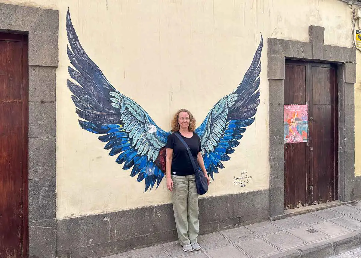 Eine Frau steht vor einem Streetart-Werk mit großen, blau-schwarzen Engelsflügeln an einer beigen Wand in Las Palmas de Gran Canaria, wodurch es so aussieht, als hätte sie selbst Flügel – ein beliebter Ort bei Besuchern, die nach Tipps zu Sehenswürdigkeiten in der Stadt suchen.