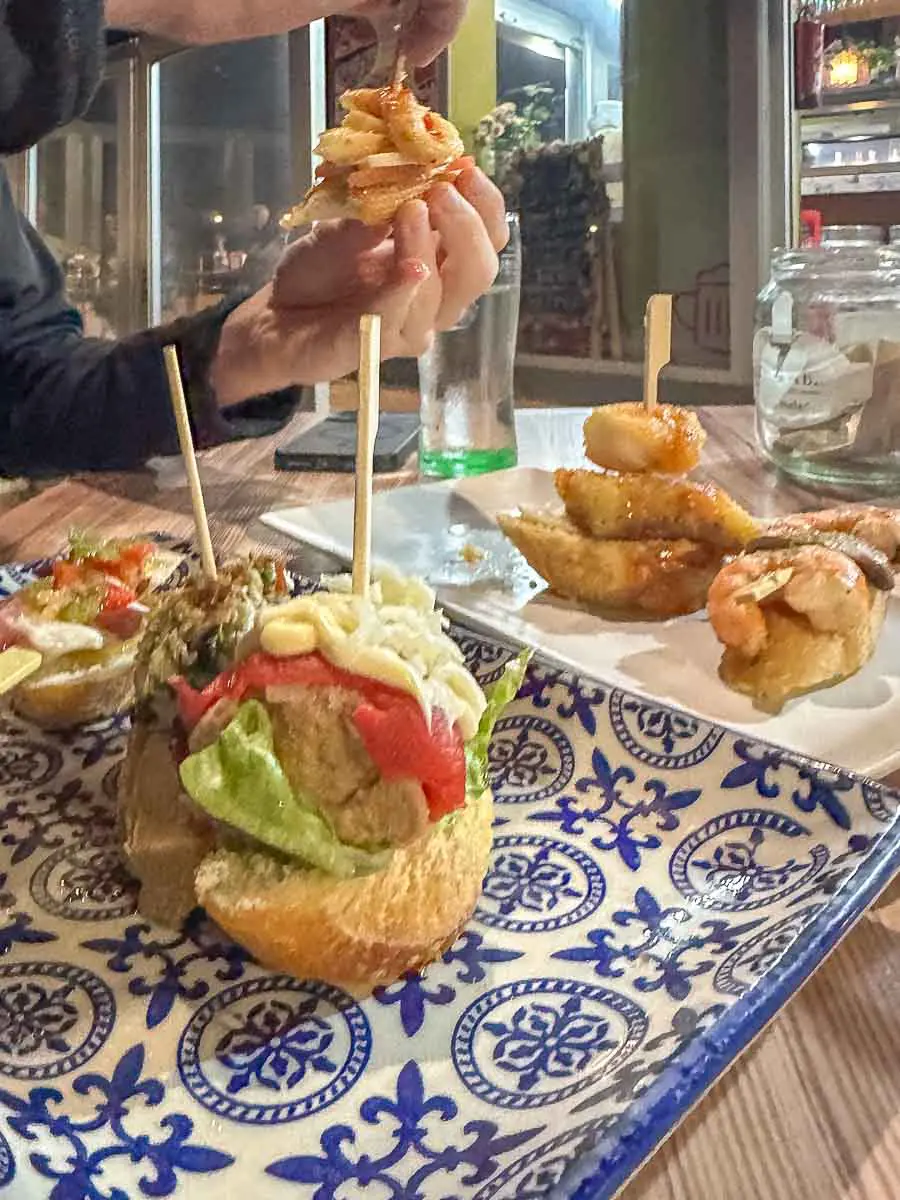Zwei Teller mit Tapas auf einem blau-weiß gemusterten Teller; auf dem einen Teller liegt Brot mit Salat, Paprika und Soße, auf dem anderen Garnelen auf Brot. Eine Hand hält einen Bissen über dem Tisch in der Luft und fängt so die intensiven Aromen ein, die man entdecken kann, wenn man Las Palmas de Gran Canaria erkundet und die lokalen kulinarischen Sehenswürdigkeiten genießt.