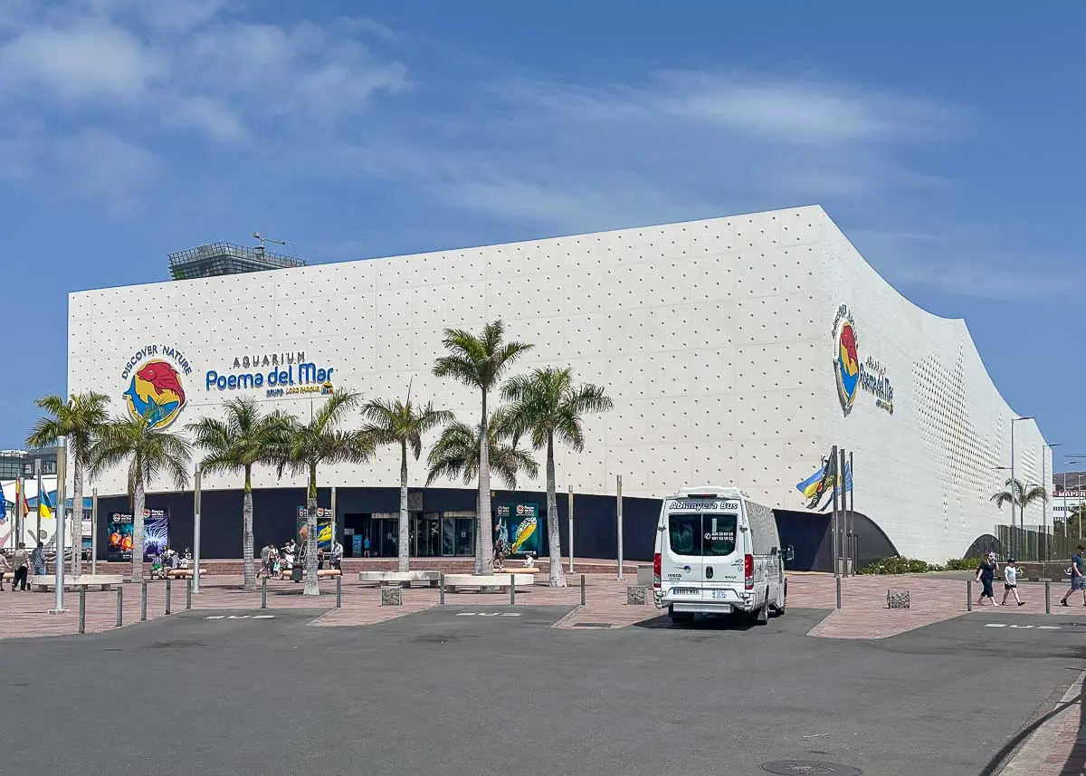 Ein großes, weißes, modernes Aquariumgebäude mit der Aufschrift „Poema del Mar“, eine der wichtigsten Sehenswürdigkeiten in Las Palmas de Gran Canaria mit Kindern, umgeben von Palmen, vorbeigehenden Menschen und einem vor dem Gebäude geparkten weißen Transporter unter strahlend blauem Himmel.