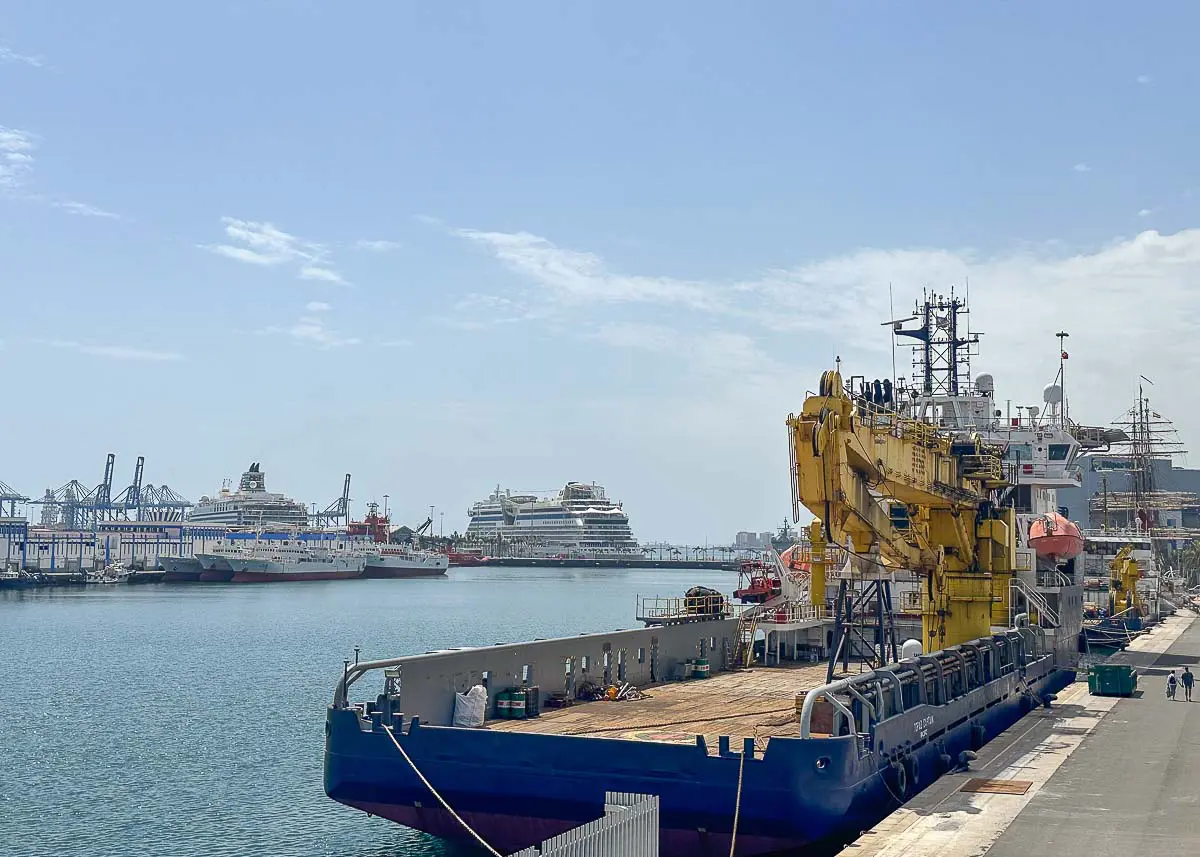 Ein vor Anker liegendes Frachtschiff mit einem gelben Kran im Vordergrund, mehrere Kreuzfahrtschiffe im Hintergrund und Industriekräne entlang der entfernten Küstenlinie unter einem teilweise bewölkten Himmel – eine typische Hafenszene, die einige der Sehenswürdigkeiten von Las Palmas de Gran Canaria zeigt.