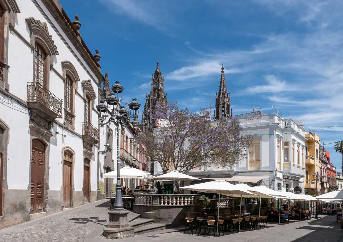 Eine lebhafte Straßenszene in der kanarischen Stadt Arucas mit Straßencafés, historischen Gebäuden, Laternenpfählen, blühenden Bäumen und Kirchtürmen unter blauem Himmel mit zarten Wolken – perfekt für alle, die authentische Erlebnisse und lebendige Momente suchen, wie sie für Las Palmas Ausflüge typisch sind.