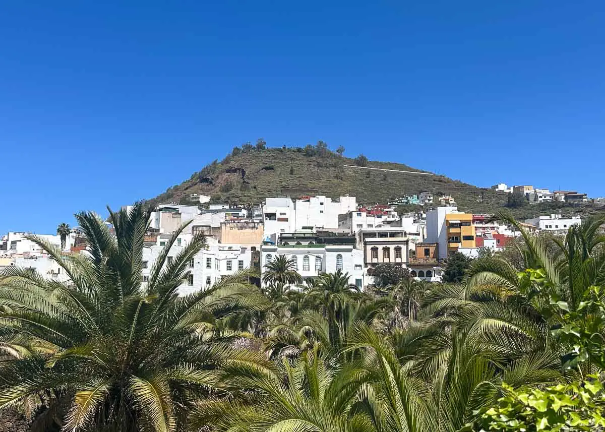 Weiße und farbenfrohe Gebäude säumen den Fuß eines grünen Hügels, umgeben von zahlreichen Palmen unter strahlend blauem Himmel – eine einladende Kulisse, die oft auf Ausflügen nach Las Palmas erkundet wird.