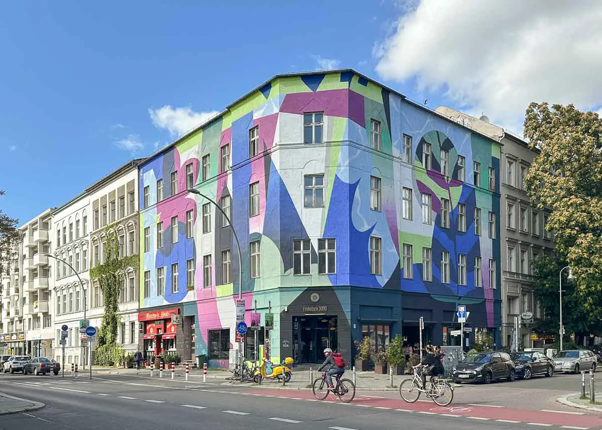 Ein farbenfrohes, geometrisches Wandbild ziert die Fassade eines Eckgebäudes gegenüber des Urban Nation Berlin an einer belebten Stadtstraße, auf der Radfahrer, Fußgänger und geparkte Autos zu sehen sind.