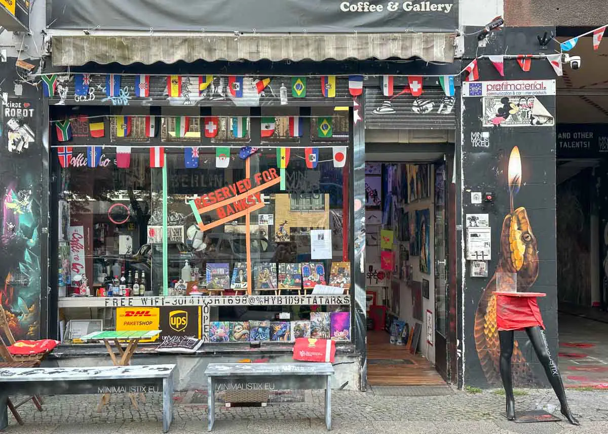 Die farbenfrohe Ladenfront von Minimalistix mit internationalen Flaggen, Büchern und Plakaten vermittelt eine lebendige Streetart Atmosphäre . Im Schaufenster hängt ein rotes Schild mit der Aufschrift „Reserved for Banksy“. Draußen stehen Bänke und eine halbe Schaufensterpuppe in einem roten Rock, die als Stehtisch dient; ein Wandgemälde mit einer Schlange ziert die Eingangswand.