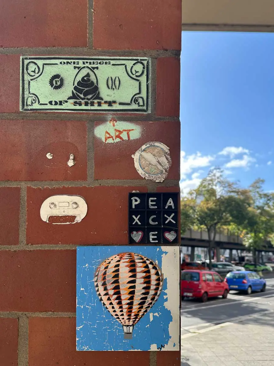 Verschiedene Street-Art-Motive an einer Backsteinmauer – darunter ein Heißluftballon, eine Kassette, ein gefälschter Dollar und Fliesen, die mit Herzen das Wort „PEACE“ bilden. Im Hintergrund sind Autos und Bäume auf der Straße zu sehen.