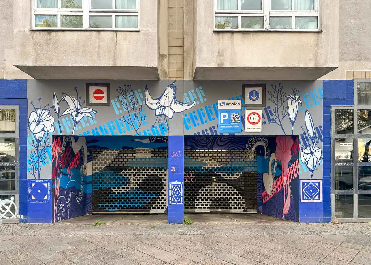 Eine Garageneinfahrt mit farbenfroher Street-Art, darunter Blumen, Vögel und Wellenmuster, unterhalb eines grauen Wohnhauses. Über der Einfahrt sind zahlreiche Park- und Verkehrsschilder angebracht.