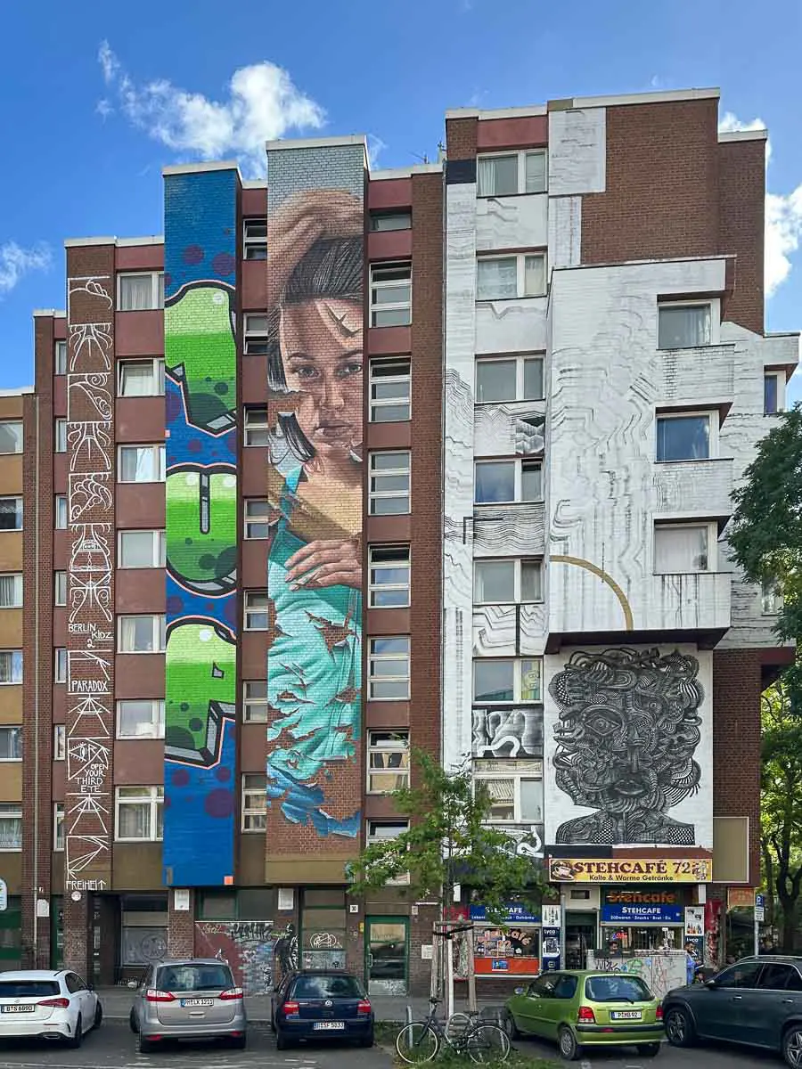 Ein Backsteinwohnhaus mit großflächigen, farbenfrohen Murals – darunter das große Porträt einer Frau, abstrakte Motive und geometrische Muster. Geparkte Autos, Fahrräder und ein Straßencafé im Erdgeschoss tragen zur lebhaften städtischen Atmosphäre bei.