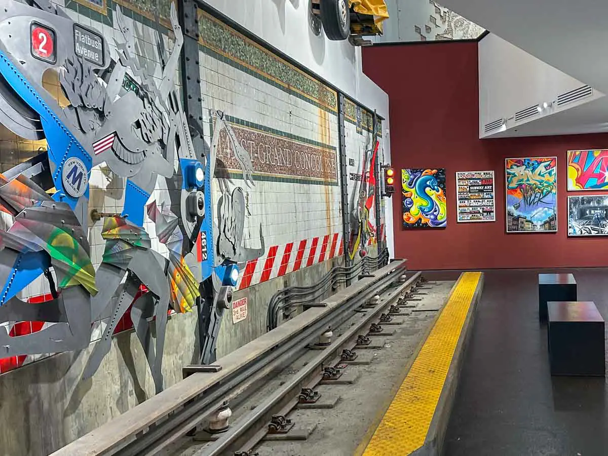 Die aktuelle Ausstellung im Urban Nation Museum (stand 2026) umfasst U-Bahn-Gleise, ein „Grand Concourse“-Schild, farbenfrohe Kunstwerke, Metallskulpturen, Bänke und lebhafte Wandmalereien im Graffiti-Stil.