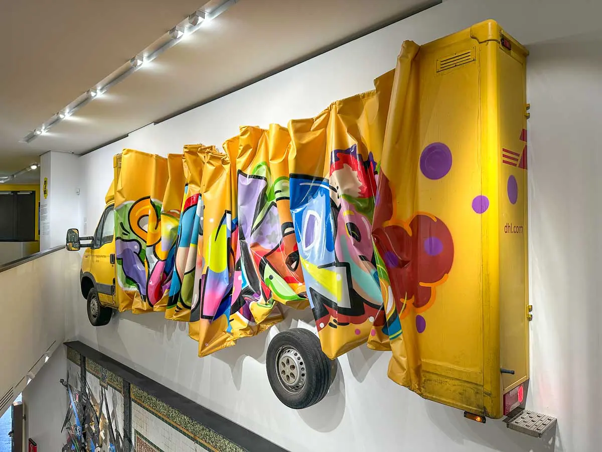 Im Urban Nation Museum ist ein gelber Lieferwagen seitlich an einer Wand befestigt, wobei sein mittlerer Teil durch eine helle, gefaltete LKW-Plane ersetzt wurde, die mit farbenfrohen Graffiti-Motiven bemalt ist. Die Installation erstreckt sich entlang einer Flurwand.