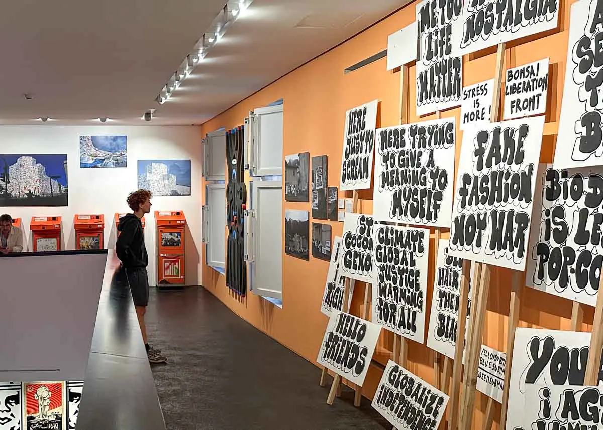 Ein Teenager betrachtet im Urban Nation Museum eine Ausstellung mit Protestschildern, auf denen fette, schwarze Schrift zu sehen ist. Die Wände sind orange, und entlang des Korridors hängen gerahmte Streetart-Kunstwerke und Plakate. Im Hintergrund ist eine weitere Person zu sehen.