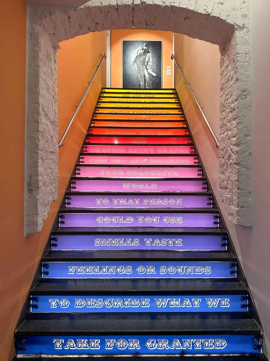 Eine Treppe im Urban Nation Museum, die in Regenbogenfarben gestrichen ist und auf deren Stufen ein inspirierendes Zitat über die Sinne zu lesen ist; oben hängt ein Schwarz-Weiß-Stencil-Kunstwerk.