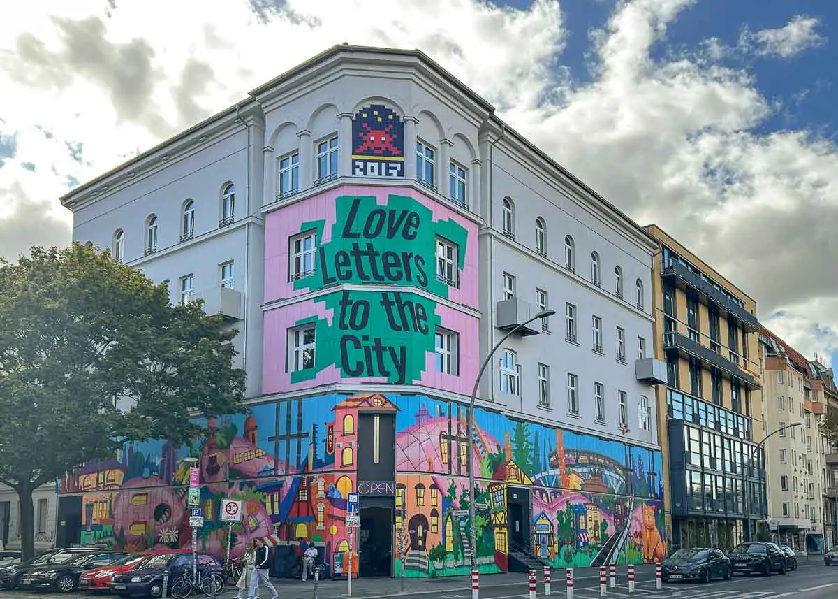 Urban-Nation-Museum-5104 An einer Straßenecke unter bewölktem Himmel steht das weiße Gebäude des Urban Nation Museum mit farbenfrohen Murals, darunter der Schriftzug „Love Letters to the City“ und lebhafte Stadtansichten.