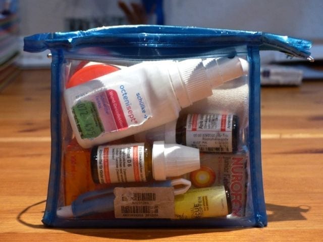 Ein transparenter, blauer Beutel mit Reißverschluss, perfekt für Familienzeit-Abenteuer, enthält verschiedene medizinische Hilfsmittel für eine Reiseapotheke. Darin befinden sich eine Wunddesinfektions-Sprühflasche, beschriftete Tropfflaschen, eine kleine Tube und eine Nurofen-für-Kinder-Schachtel. Der Beutel liegt auf einer Holzoberfläche, im Hintergrund sind unscharfe Papiere zu sehen.