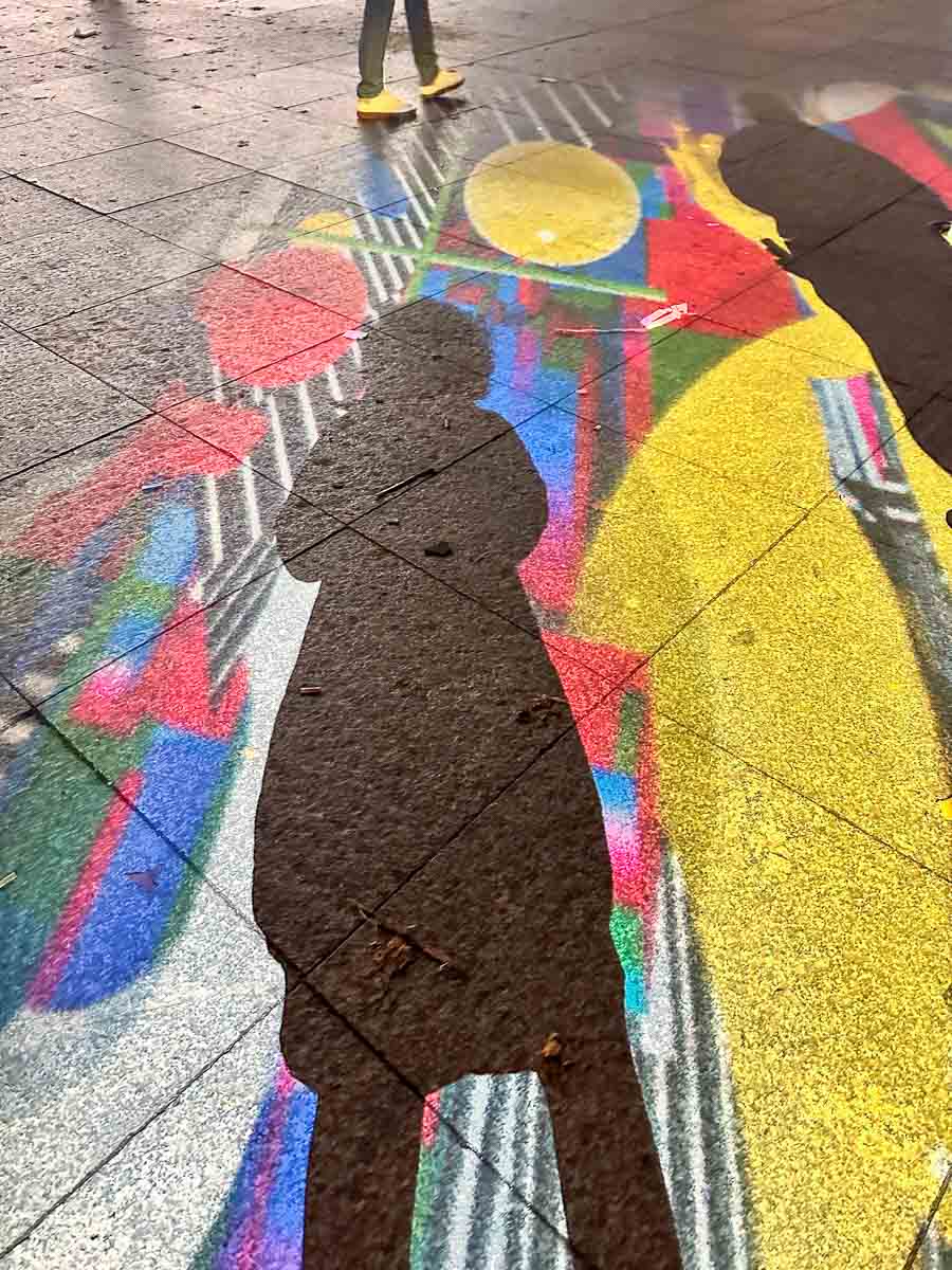 Der Schatten einer Person fällt auf eine bunte Kreidezeichnung von Menschen auf dem nassen Bürgersteig und fängt so die verspielte Stimmung ein, die beim „Festival of Lights mit Kindern“ oft zu spüren ist. Eine andere Person, die gelbe Schuhe trägt, geht in der Nähe vorbei.