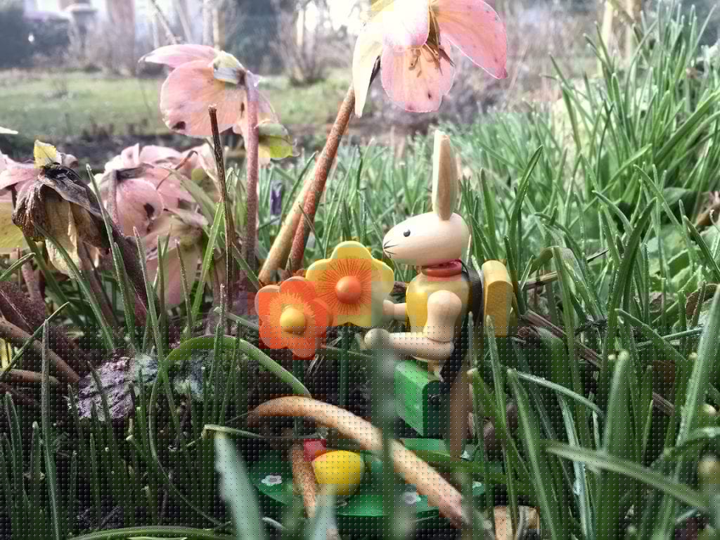 Eine hölzerne Hasenfigur sitzt auf grünem Gras, umgeben von rosa Blumen und Pflanzen. Sie hält eine kleine Gießkanne und steht neben einer Gruppe orangefarbener und gelber Blüten, was eine skurrile Osterstimmung in Berlin hervorruft. Die Szene wirkt zugleich skurril und heiter.