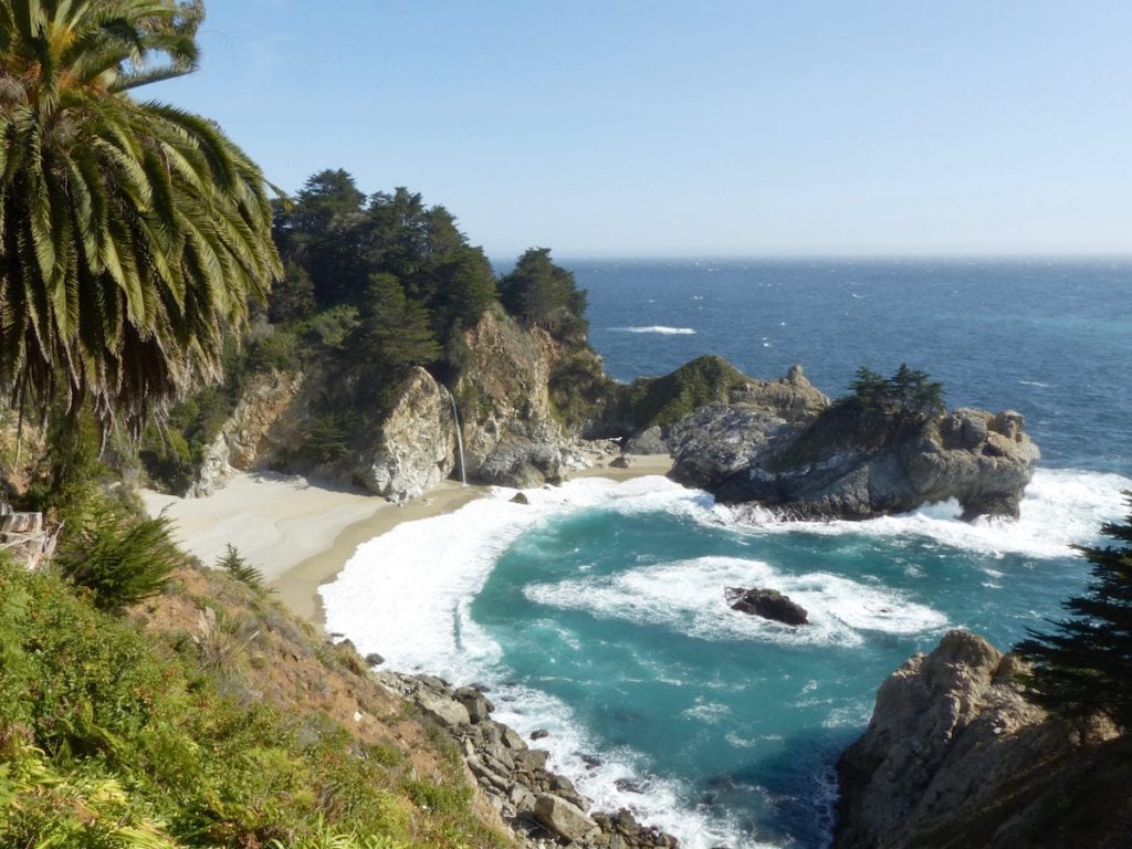 Ein Blick auf die Küste am Highway 1 offenbart einen Strand im Julia Pfeiffer Burns State Park mit türkisfarbenem Wasser und weißen Wellen, die an das Sandufer schlagen, eingerahmt von felsigen Klippen, von denen ein Wasserfall rauscht und üppigem Grün, darunter hohe Palmen unter einem klaren blauen Himmel.
