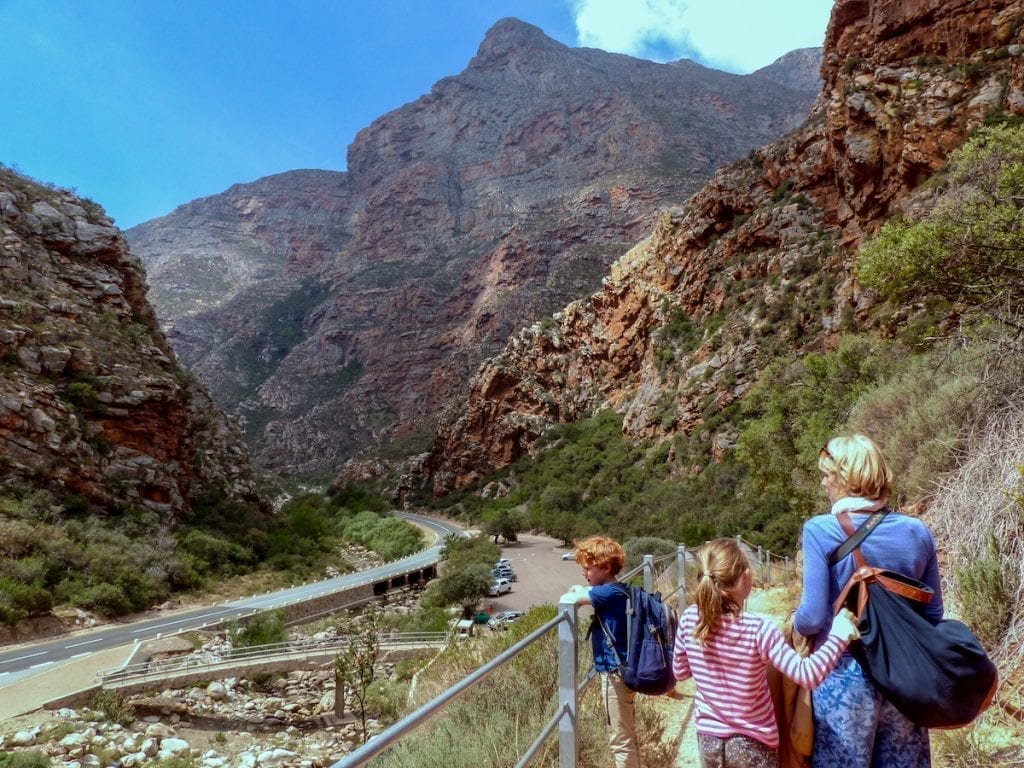 Eine Familie wandert auf einem geländergesäumten Pfad durch eine felsige Bergregion. Die Eltern und zwei Kinder genießen die malerische Aussicht, die an „Südafrika mit Kindern“ erinnert. Unten ist eine kurvenreiche Straße mit parkenden Autos zu sehen, im Hintergrund ein hoch aufragender Berg unter einem strahlend blauen Himmel.