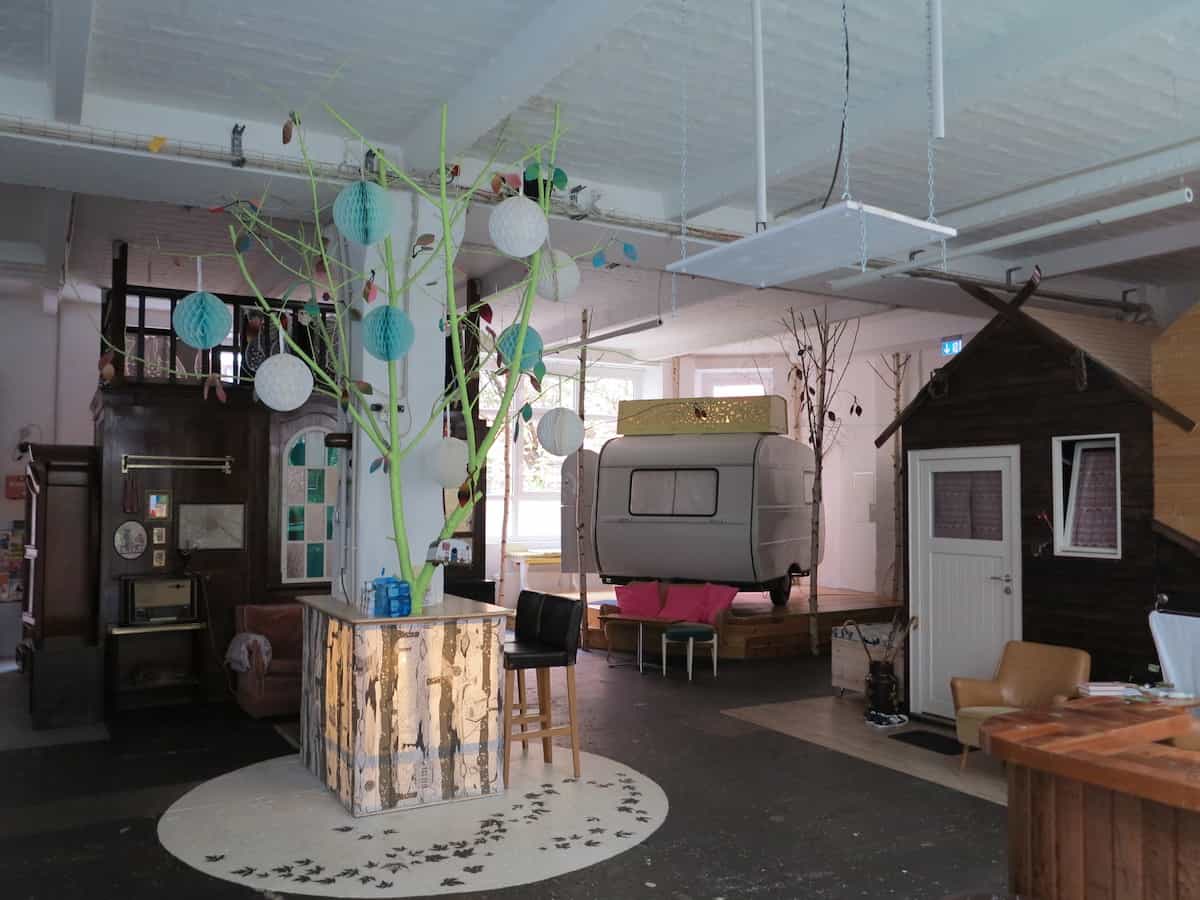 Ein großzügiger, kreativ gestalteter Büroraum mit einem über dem Boden schwebenden Wohnwagen bietet die perfekte Atmosphäre für eine ungewöhnliche Übernachtung in Berlin – komplett mit einem kleinen Holzhäuschen, einem dekorativen Baum mit Papierlaternen und einer eklektischen Einrichtung.