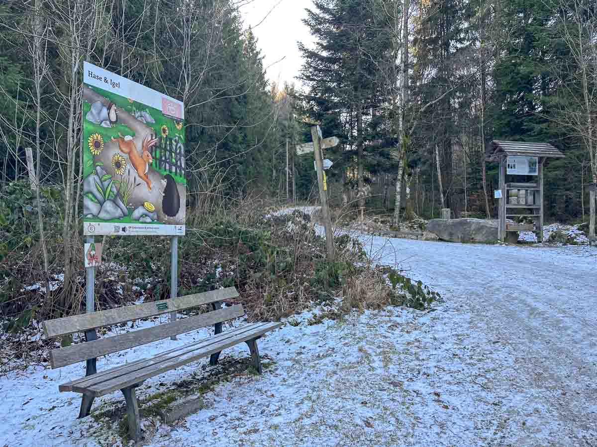 Ein verschneiter Waldweg im Fichtelgebirge lädt im Winter mit Kindern zum Spielen ein: Eine gemütliche Holzbank, ein fröhliches Schild mit Cartoon-Hase und Igel, hohe Bäume, hilfreiche Wegweiser und eine Informationstafel machen den Familienausflug zauberhaft.