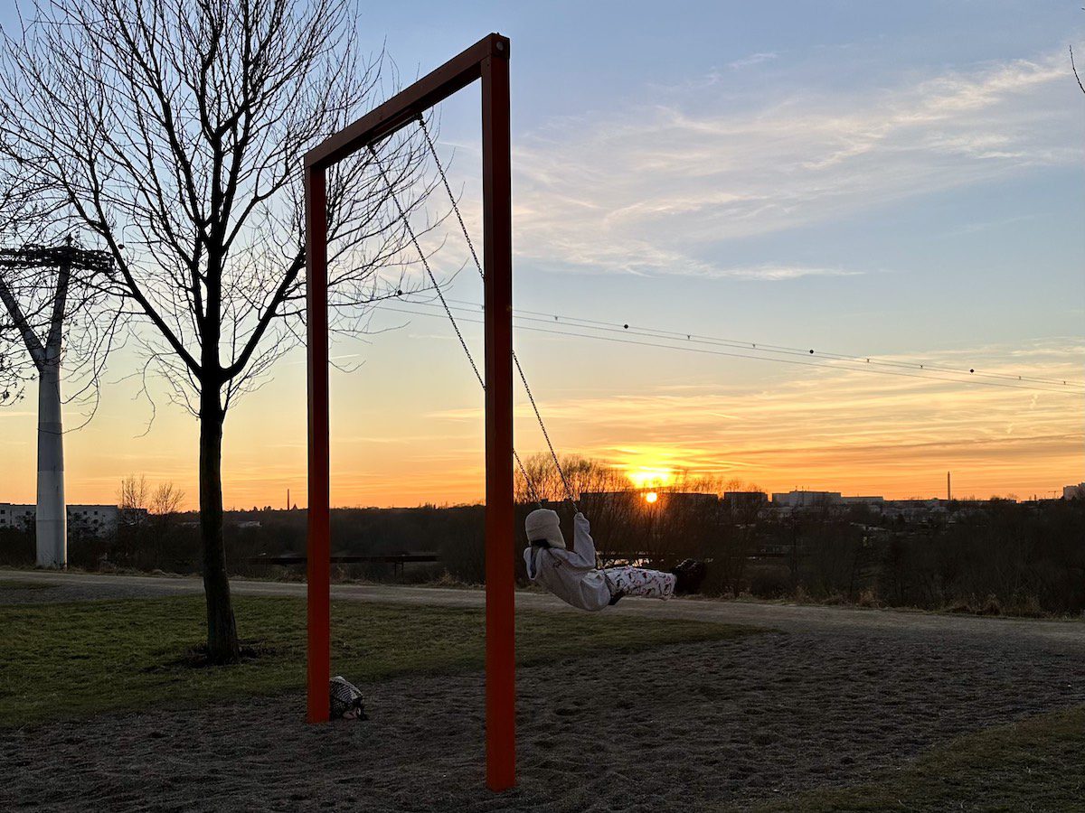 Ein Kind schaukelt auf einem Aussichtsberg in Berlin im Frühling unter kahlen Bäumen auf der grünen Wiese bei Sonnenuntergang auf einer großen Schaukel. 