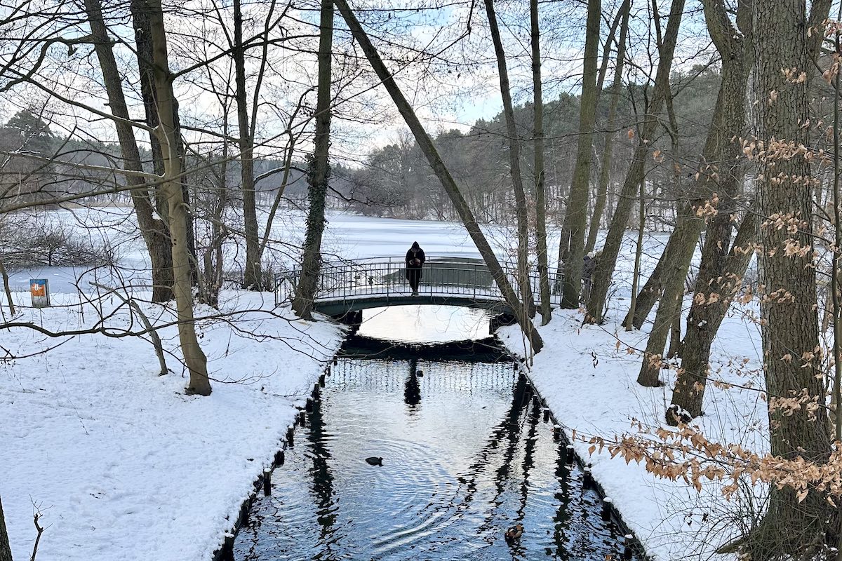 In einer verschneiten Winterlandschaft mit Wald fließt ein Kanal, in dem sich der blaue Himmel spiegelt in einen See. Auf einer Brücke steht ein Mensch.
