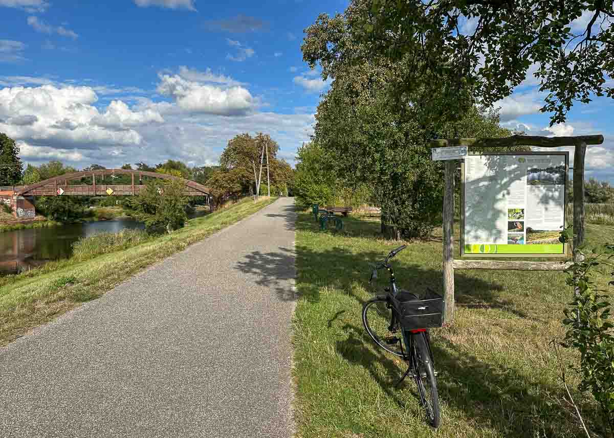 Ein Radweg verläuft entlang eines Kanals. In der Ferne ist eine hölzerne Brücke zu sehen. Unter einem Baum steht ein Fahrrad neben einer Informationstafel. Der Himmel ist blau mit Wölkchen.