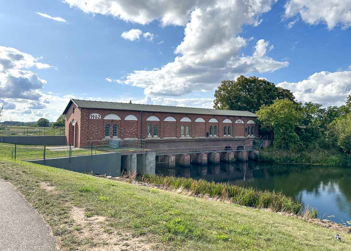 Ein historisches Backsteingebäude mit Bogenfenstern steht am Ufer eines ruhigen Flusses unter einem teilweise bewölkten Himmel im Herzen des Nationalparks Unteres Odertal. Üppig grüne Bäume und Gras umgeben das Gelände, und auf dem Gebäude ist die Jahreszahl 1907 zu sehen.