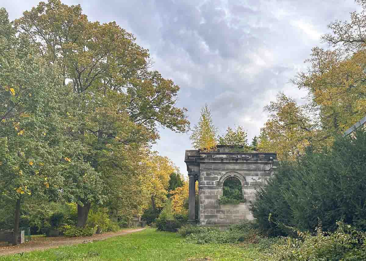 Eine steinerne Ruine mit einem Bogenfenster steht am Rande eines grasbewachsenen Weges auf einem der historischen Friedhöfe in Berlin, umgeben von grünen und herbstlichen Bäumen unter einem wolkenverhangenen Himmel