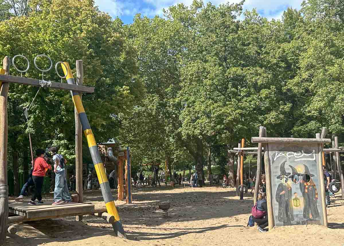 Kinder spielen in einem sonnigen Park mit Klettergeräten und einer Maltafel. Hohe grüne Bäume spenden Schatten, und im Hintergrund sind mehrere Menschen zu sehen, die die freie Natur genießen. Wie viele Parks in Berlin bietet auch der Volkspark Wilmersdorf Familien einen lebendigen und einladenden Ort, um sich zu entspannen und Spaß im Freien zu haben.
