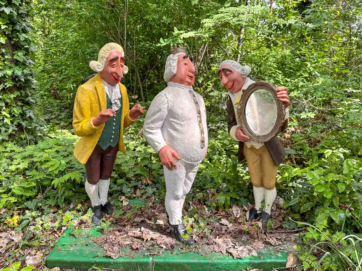 Drei Statuen von Männern in altmodischer Kleidung stehen inmitten von Grün im Märchenhain der Gärten der Welt Berlin. Sie zeigen eine Szene aus dem Märchen "Des Kaisers neue Kleider". Einer trägt Gelb, einer Weiß und einer Braun, während er einen runden Spiegel in der Hand hält - eines der Highlights in einem der berühmtesten Parks in Berlin.