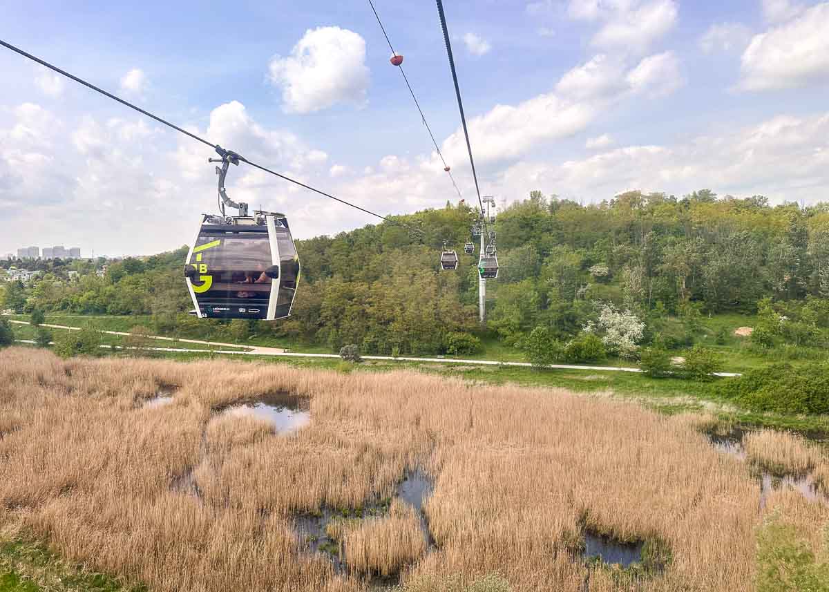 Seilbahnen fahren über das grasbewachsene, sumpfige Wuhletal hinweg, im Hintergrund sind ein bewaldeter Hügel und der blaue Himmel zu sehen – ein Bild, das die landschaftliche Schönheit widerspiegelt, wie man sie oft in Berliner Parks vorfindet.