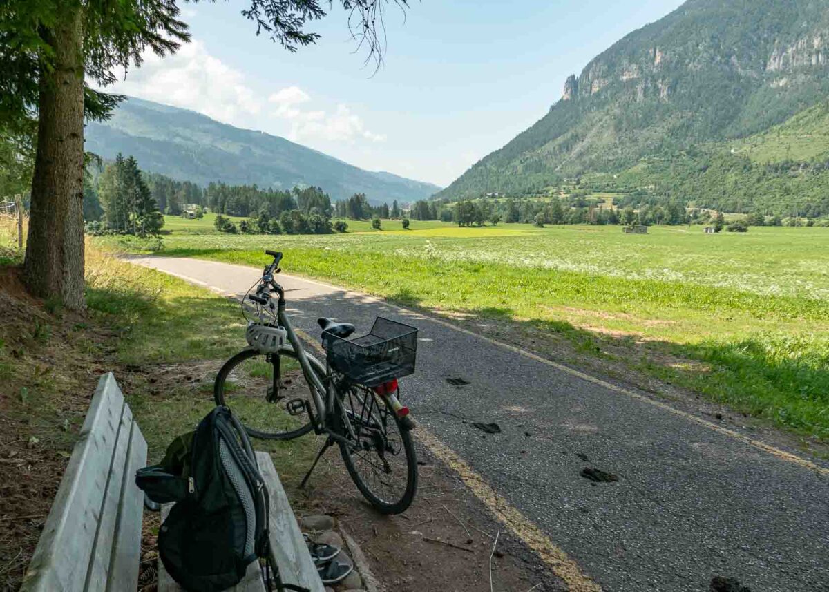 Geteerter Radweg in den Bergen mit Fahrrad an einer schattigen Rastbank 