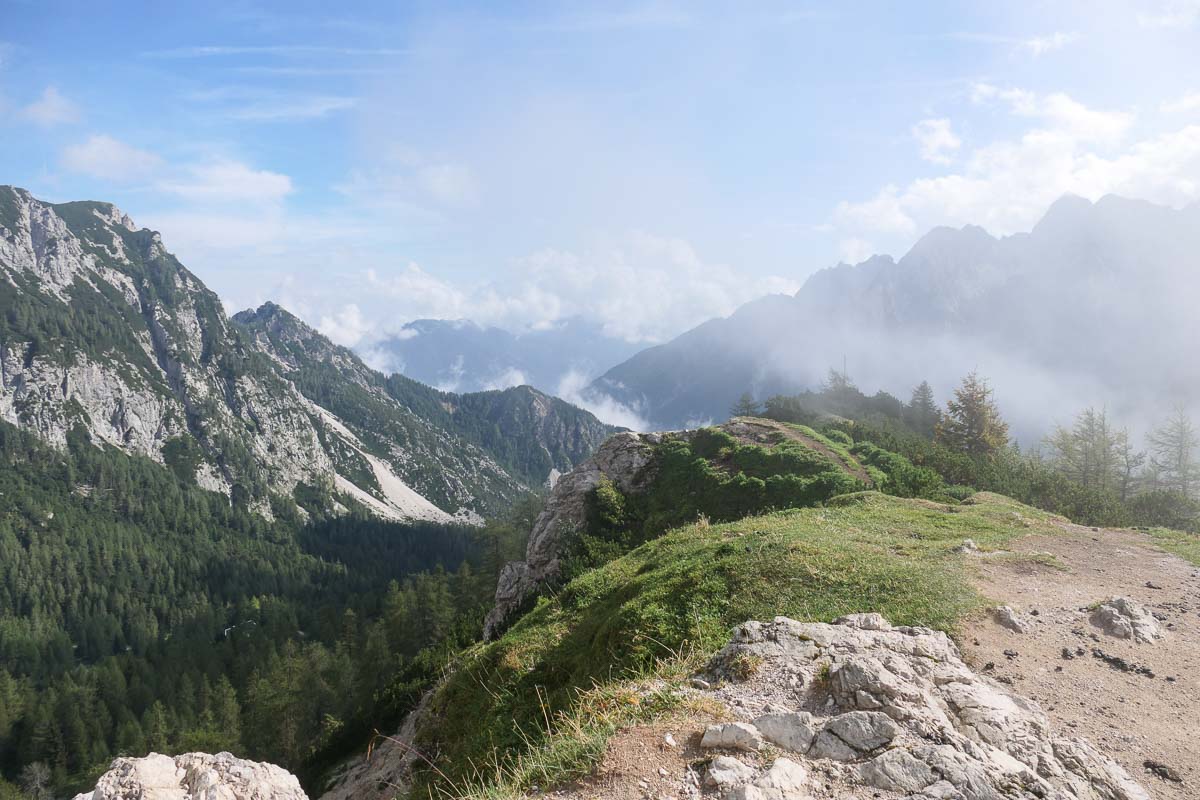 Atemberaubende Berglandschaft mit Wolkenfetzen zwischen Felsengipfeln und bewaldeten Berghängen. Im Vordergrund sieht man eine kahle Bergkuppe.